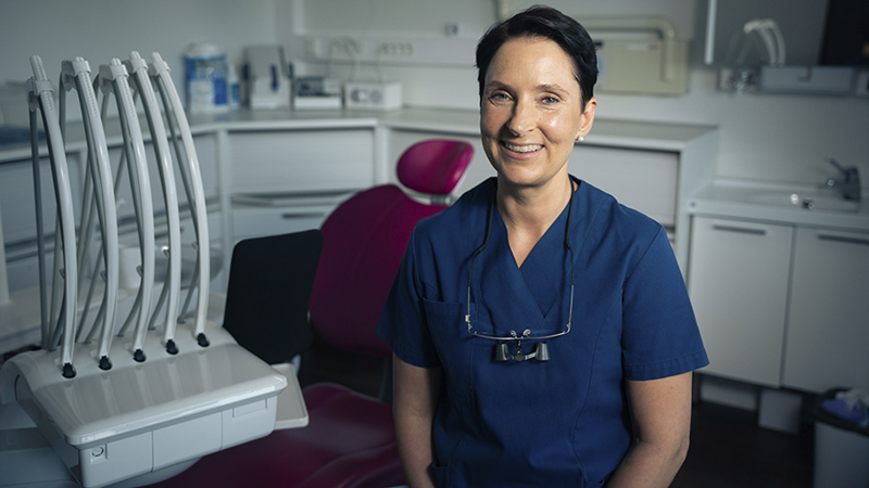 Hammaslääkäri istuu hoitotuolin edessä ja hymyilee kameralle